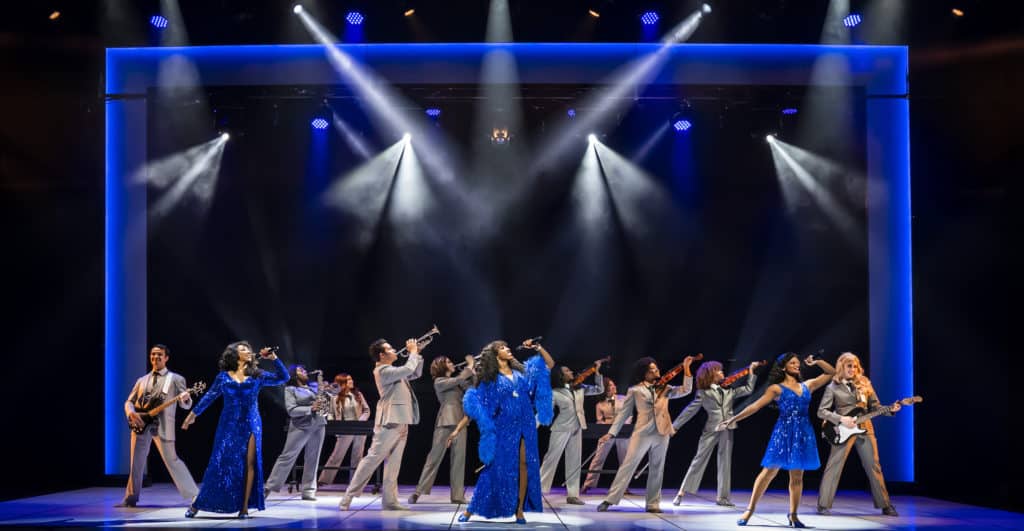 Brittny Smith as “Diva Donna,” Charis Gullage as “Disco Donna” and Amahri Edwards-Jones as “Duckling Donna" are standing on stage, looking up as they sing in front of the ensemble. 