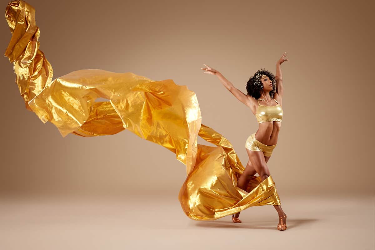 A woman in a gold costume dances with her arms up. Her costume has a gold, flowing trail that is waving in the air.
