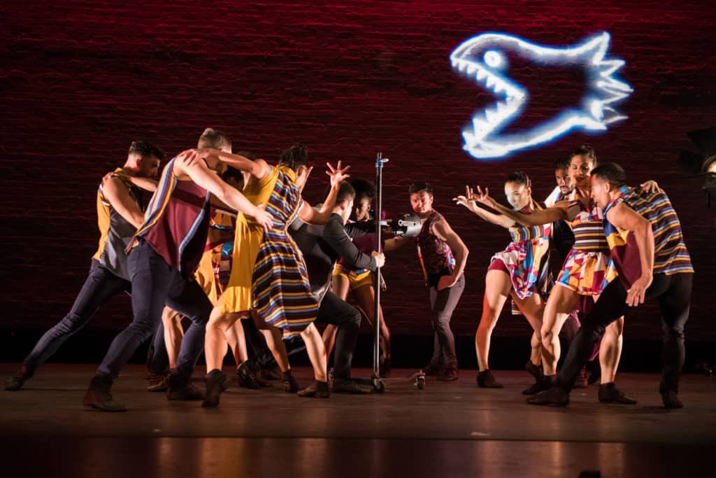 The cast of Ballet Hispánico dances on stage with a brick wall behind them. They are dancing around a stage light, and there is a neon shark figure on the wall.