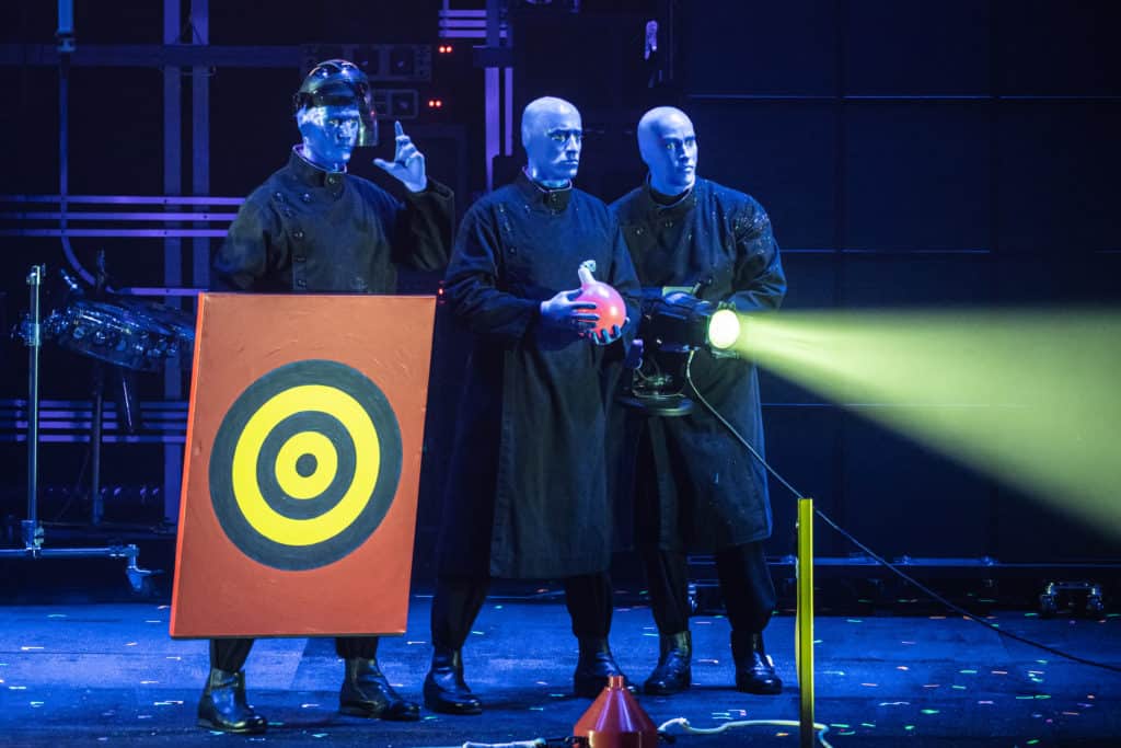The three blue men hold objects on stage including a target, a bulbous object and a spotlight. They are all wearing black smocks on stage.