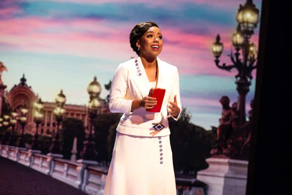 Anastasia (Kyla Stone) is looking into the distance as she sings, holding a red purse in her hand. She's wearing a white outfit, with her hair pulled up.