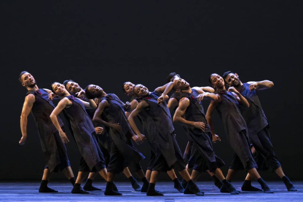The cast of Ballet Hispánico dances on stage wearing all black costumes with black socks. They are dancing in a row and leaning back as they dance.