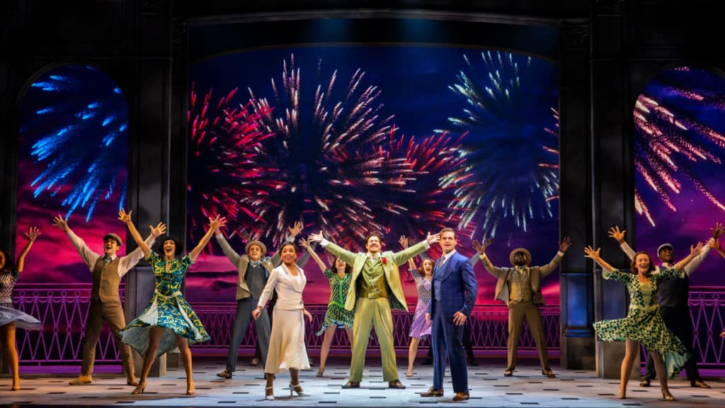 The company of Anastasia is standing in front of a firework backdrop. They're looking out to the audience with their arms up in excitement as they sing.