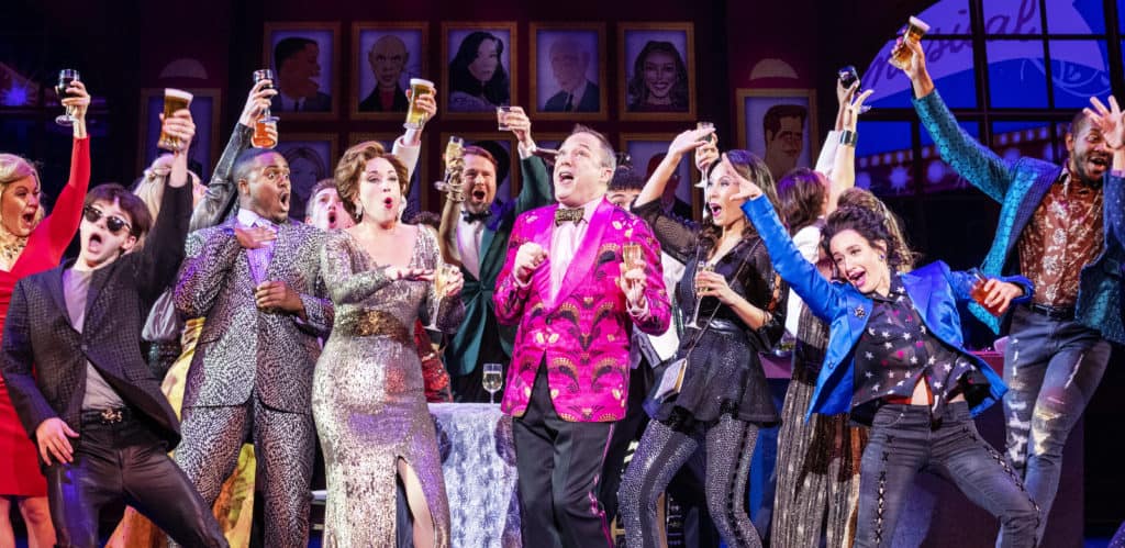 The cast of The Prom are celebrating together, holding drinks up as they cheer with one another in nice clothing.