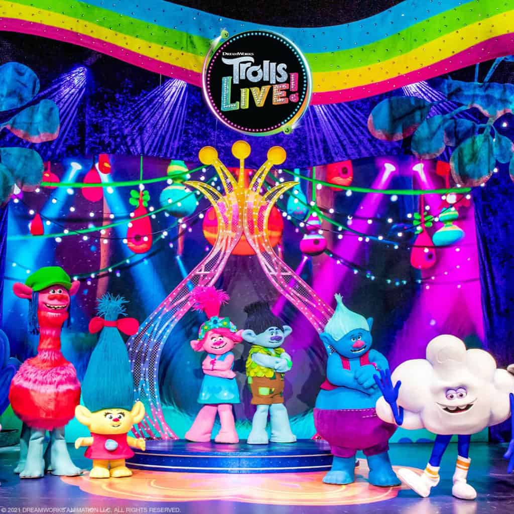 Trolls characters Cooper, Smidge, Poppy, Branch, Biggie, and Cloud Guy stand on the stage. A Trolls LIVE! logo is at the top center with rainbow streaks waving behind it. The stage and background are full of bright colors and lights.