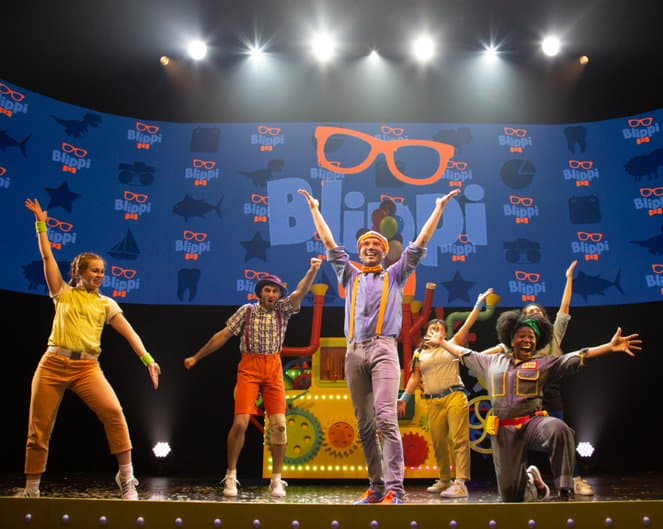 The cast of Blippi The Musical performs on stage. They all have their hands up in the air and are wearing bright, colorful outfits. The backdrop says "Blippi" and there is colorful decorations on stage
