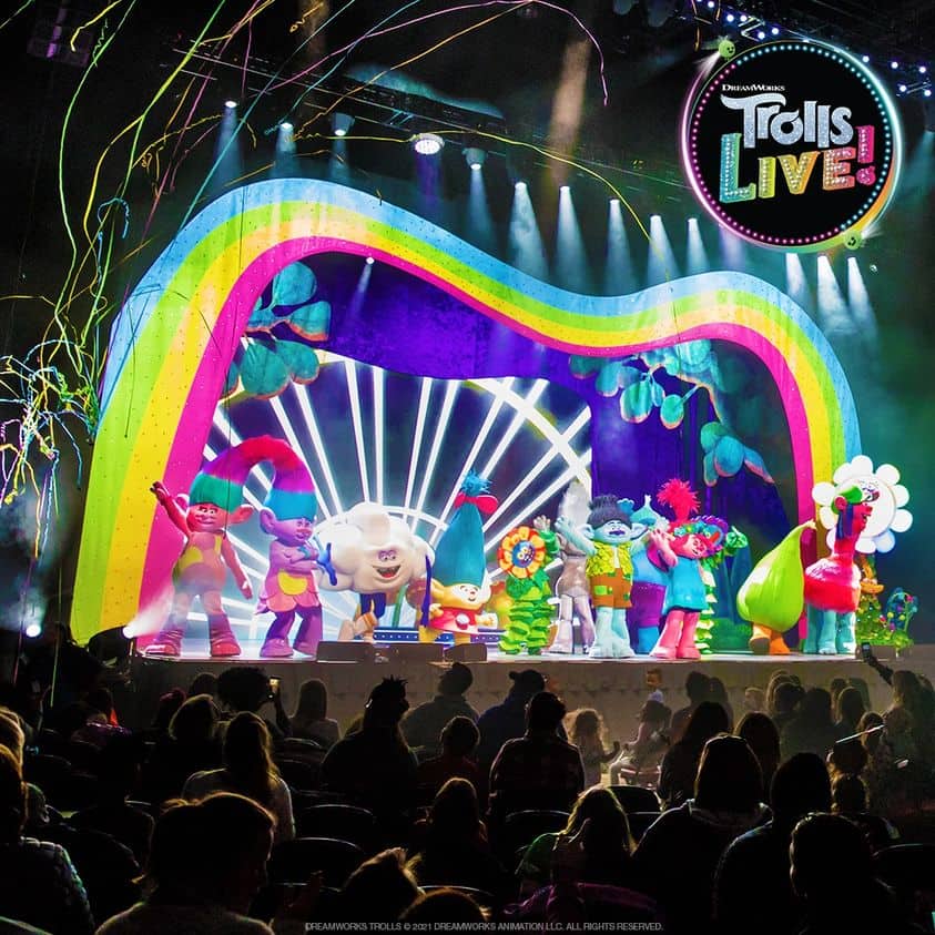 Trolls characters stand side by side on the stage, waving to the audience. A Trolls LIVE! logo is at the top right with rainbow streaks waving behind it. The stage and background are full of bright colors and lights.