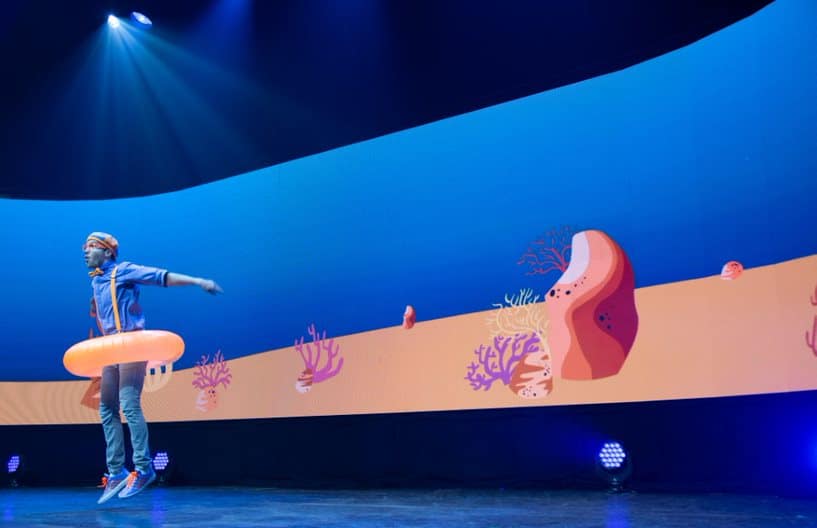 Blippi from Blippi The Musical dances on stage wearing a pool float tube around his waist. The background behind him is themed to look like underwater and the ocean.