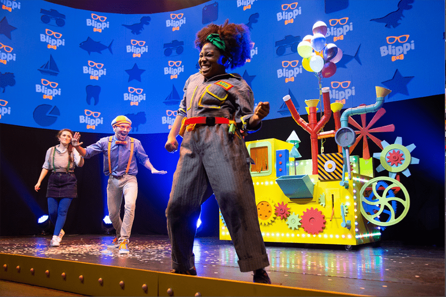 The cast of Blippi The Musical dances around on stage. A performer in a striped jumpsuit is dancing closest to the camera, with Blippi and another cast member dancing in the background. The "Blippi" logo fills the screen in the background and colorful decorations are on the stage behind the performers.