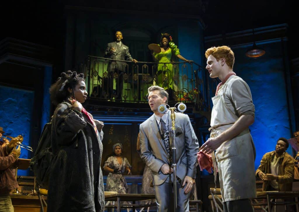 Three characters from the cast of Hadestown  stand center stage with people and instruments gathered around them. Behind them on a balcony are two people looking down at them. 