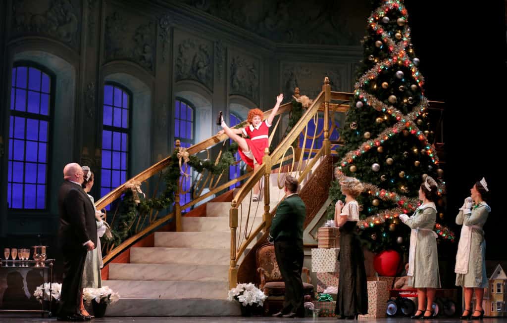 Annie walks down a staircase wearing a red dress, and kicks her leg in the air. The set is decorated for Christmas and there is a giant Christmas tree next to the stairs. Her family waits at the bottom of the stairs.
