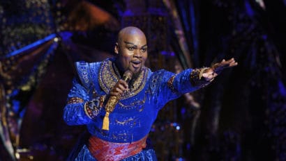 Michael James Scott as The Genie in Aladdin. He is singing and looking out to the audience, with his left arm reaching out. His outfit is blue and he has a red belt.