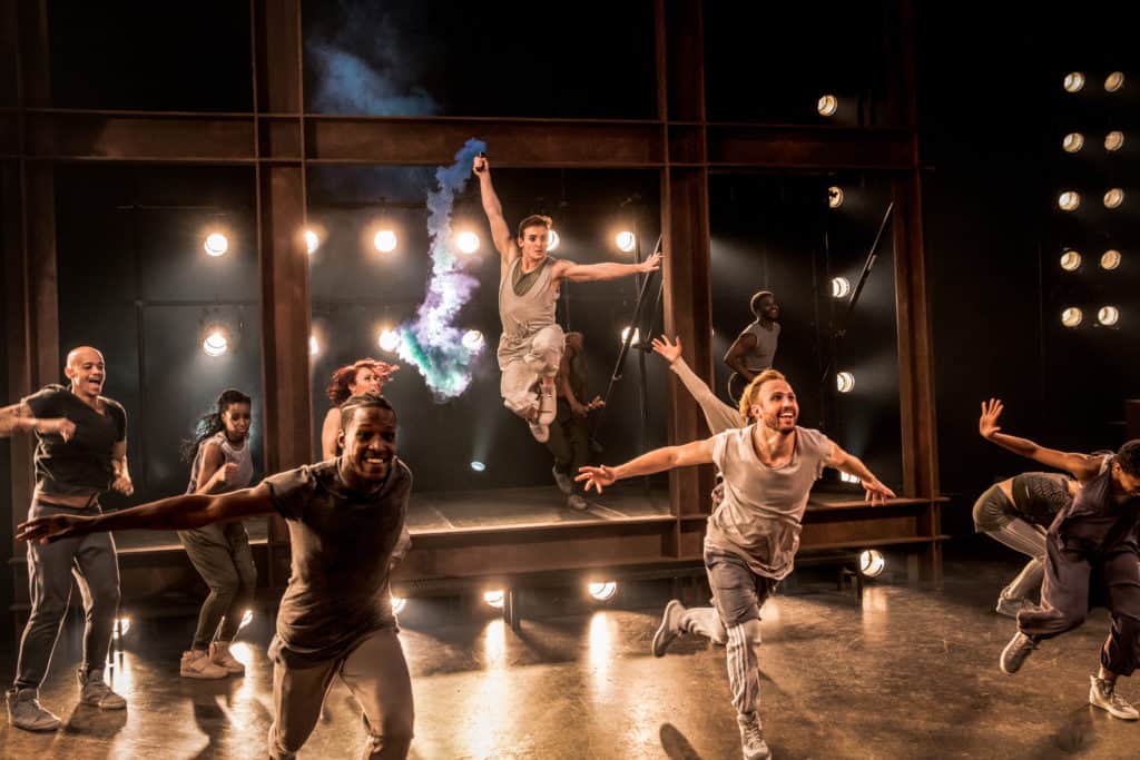 The cast of Jesus Christ Superstar dances on stage. One dancer centerstage is holding a device that is releasing colorful smoke as they dance. There are spotlights coming from behind them and the backdrop is black.
