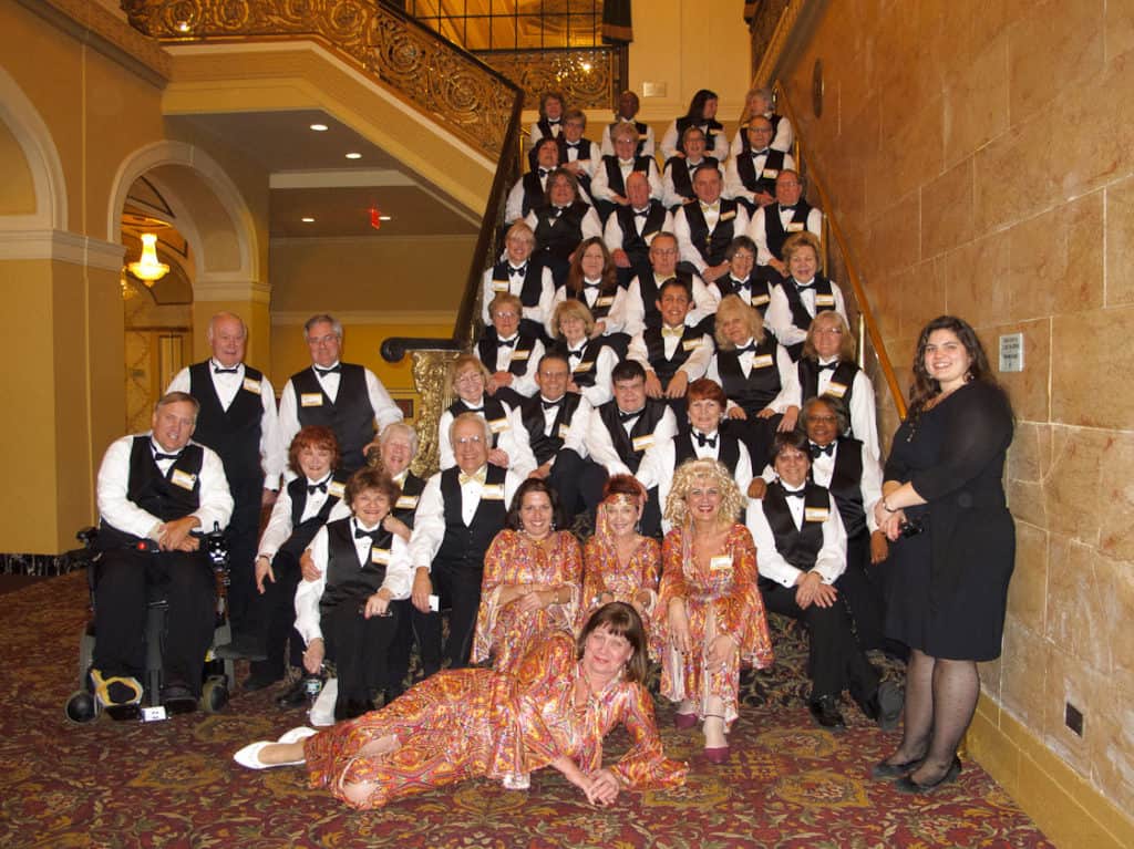 The Hanover Theatre's volunteers sit on the grand staircase. Four volunteers in the front of the group are dressed in 70's outfits. The rest of the volunteers are dressed in a black vest and tie.