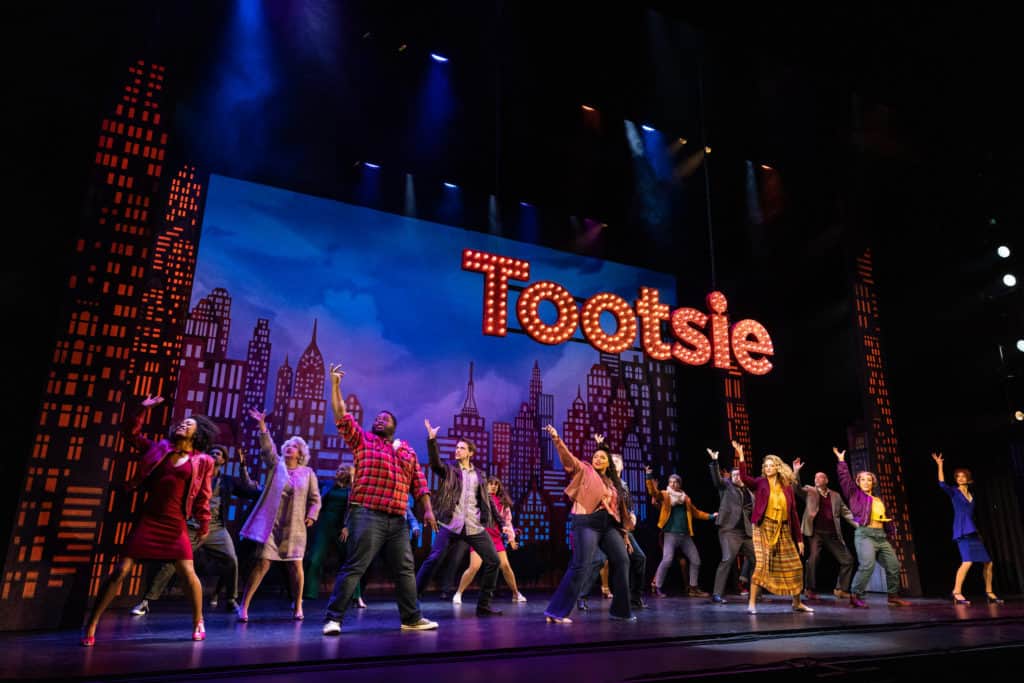 The cast of Tootsie dances on stage. Behind them, a city skyline is on the wall and a big light-up sign that says "Tootsie." The cast all has their right arm raised.