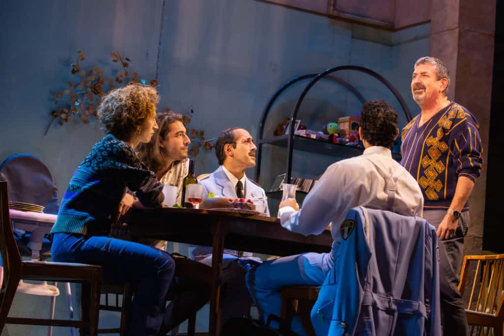 The cast of The Band's Visit performs on stage. 4 people sit around a table and one man stands at the head of the table. There is a wine bottle on the table and everyone is looking up at the man standing.