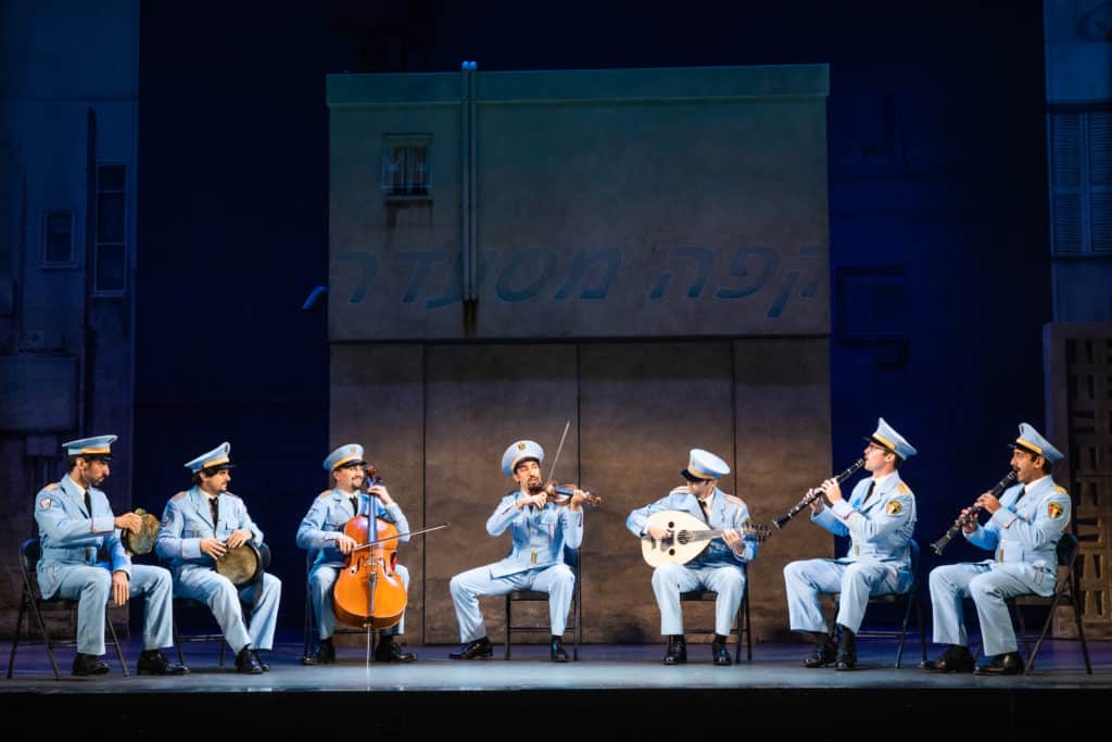 The cast of The Band's Visit performs on stage. The Alexandria ceremonial police orchestra wears blue uniforms and sit in a row playing instruments. 