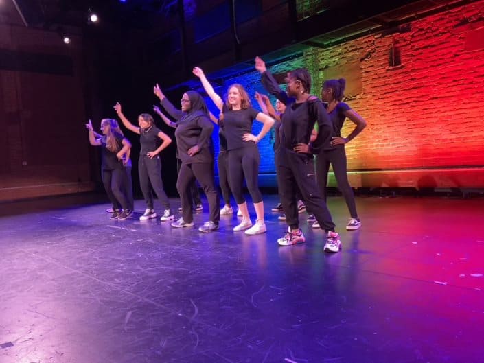 WYSH Program students wearing all black perform on stage at the BrickBox with a rainbow background behind them.