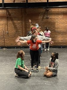 WYSH students are standing in a line on the BrickBox stage as they rehearse this year's show. Two students are sitting on either side of the line of students, looking up at the person in the front. As the front student moves their arms, the students behind are copying them.