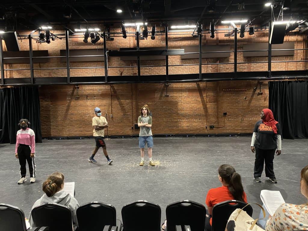 WYSH students are standing on the BrickBox stage as they rehearse this year's show. They're dressed casually, looking to each other as they move through a scene.