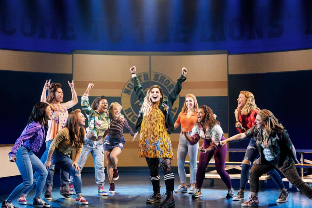 Lindsay Heather Pearce, playing Janis Sarkisian, stands in the center of the cast with her arms raised in triumph. She is wearing a yellow dress, forest green jacket and black boots with black knee-high socks. The cast around her cheers her on. 