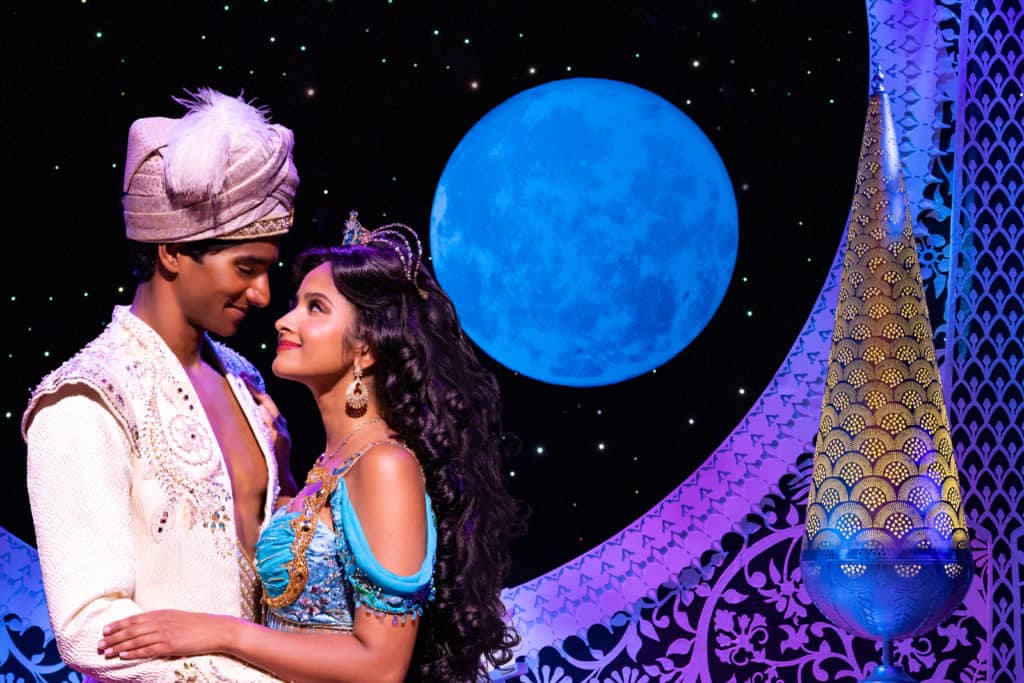 Michael Maliakel, playing Aladdin, and Shoba Narayan, playing Jasmine embrace in a hug while looking into each other's eyes. Jasmine is wearing a teal blue top with a tiara. Aladdin is wearing a white tunic and a turban. In the background is the night sky with a blue moon and ornate decorations. 