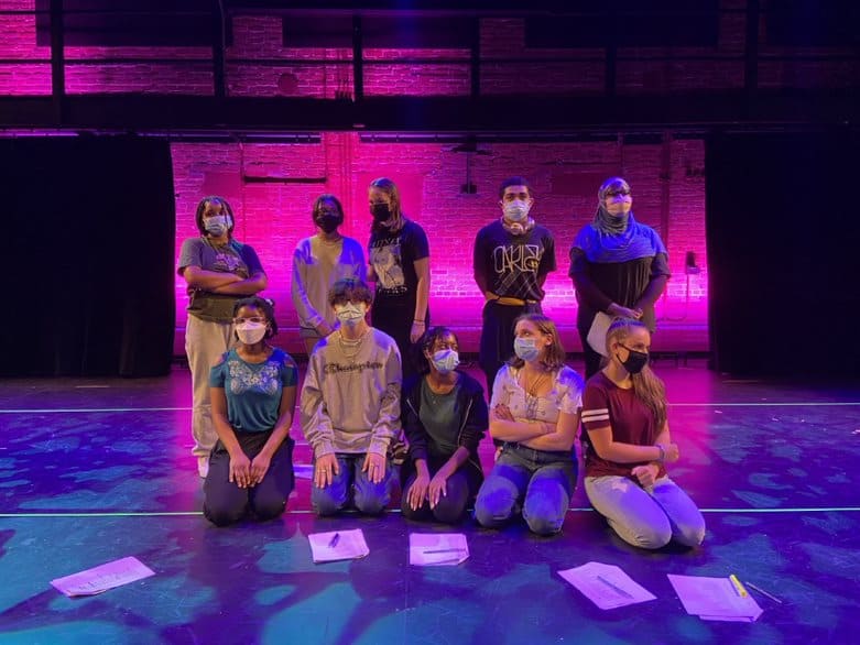 WYSH Program students pose on stage at the BrickBox. A pink background illuminates the bricks behind them, and the stage has a blue glow. The students are wearing masks.