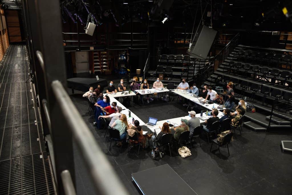 The cast of Macbeth sits at the BrickBox for the script reading. They sit with 4 white tables in a square, some turned around to look at something off screen.