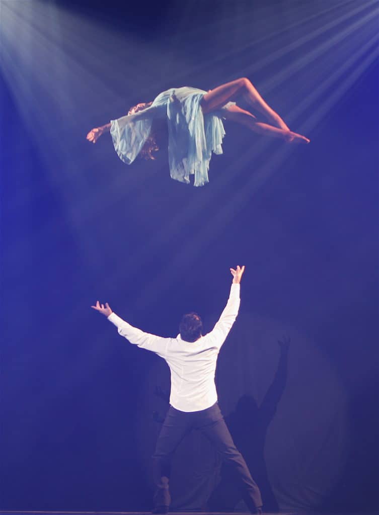A magician is seen making a woman levitate. She is wearing a blue dress and is floating horizontally on stage.