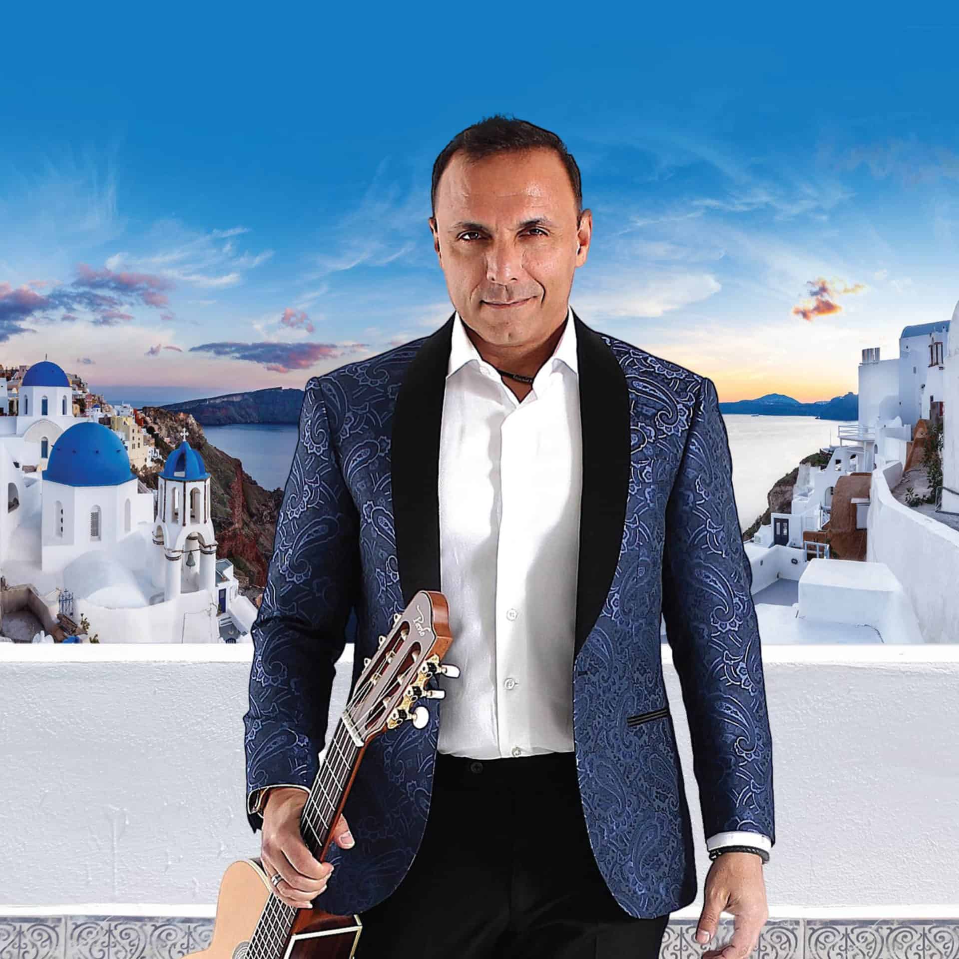 Pavlo standing in front of Greece, white houses and a blue sky behind him. He is holding a guitar in his hand and wearing a blue jacket.