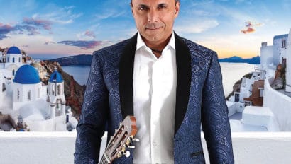 Pavlo standing in front of Greece, white houses and a blue sky behind him. He is holding a guitar in his hand and wearing a blue jacket.