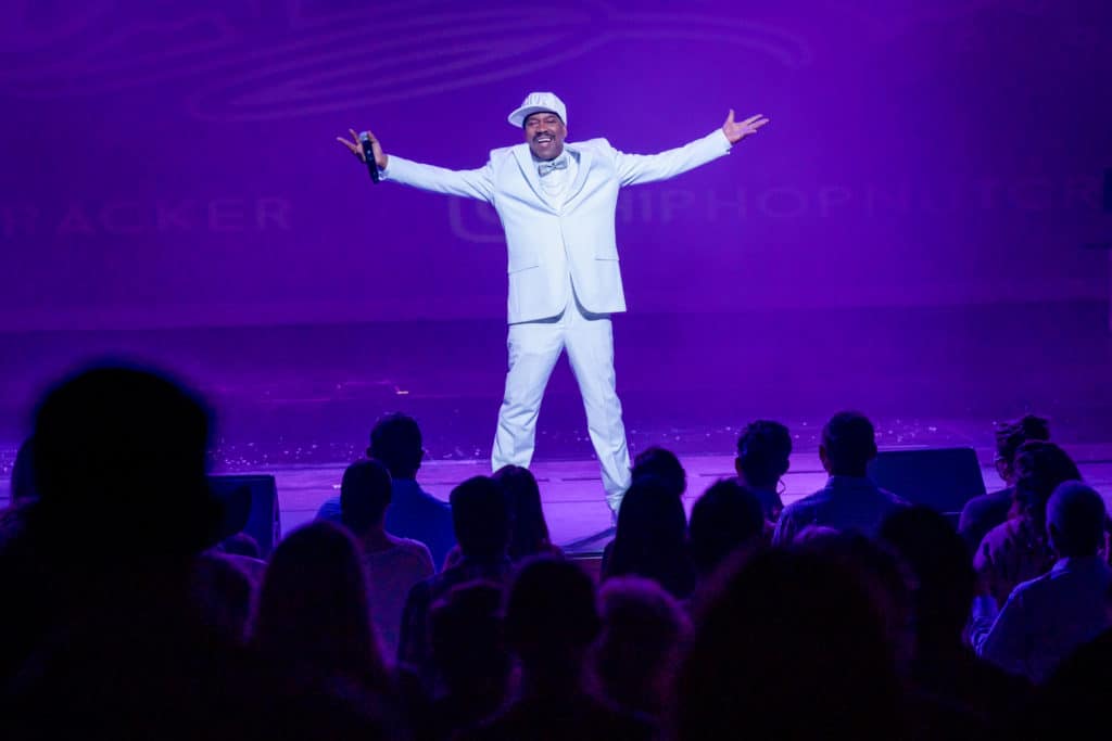 Kurtis Blow stands on stage in front of a purple background. He wears a full white suit and white hat. He stands with his arms extended and holds a microphone in his right hand.