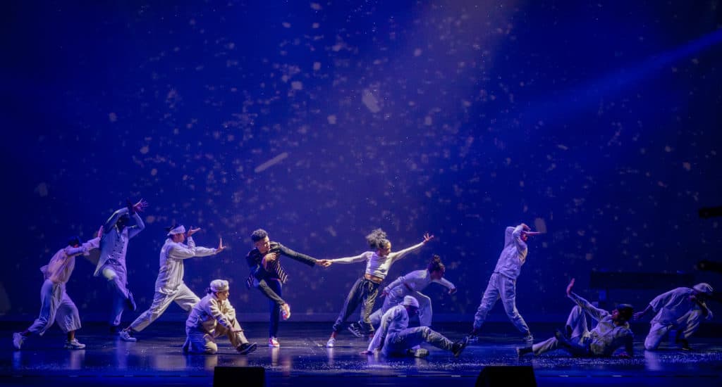 The company of Hip Hop Nutcracker dances on stage in front of a purple background. They are dancing and there are two people holding hands in the center of the stage. Everyone around the couple is wearing white.