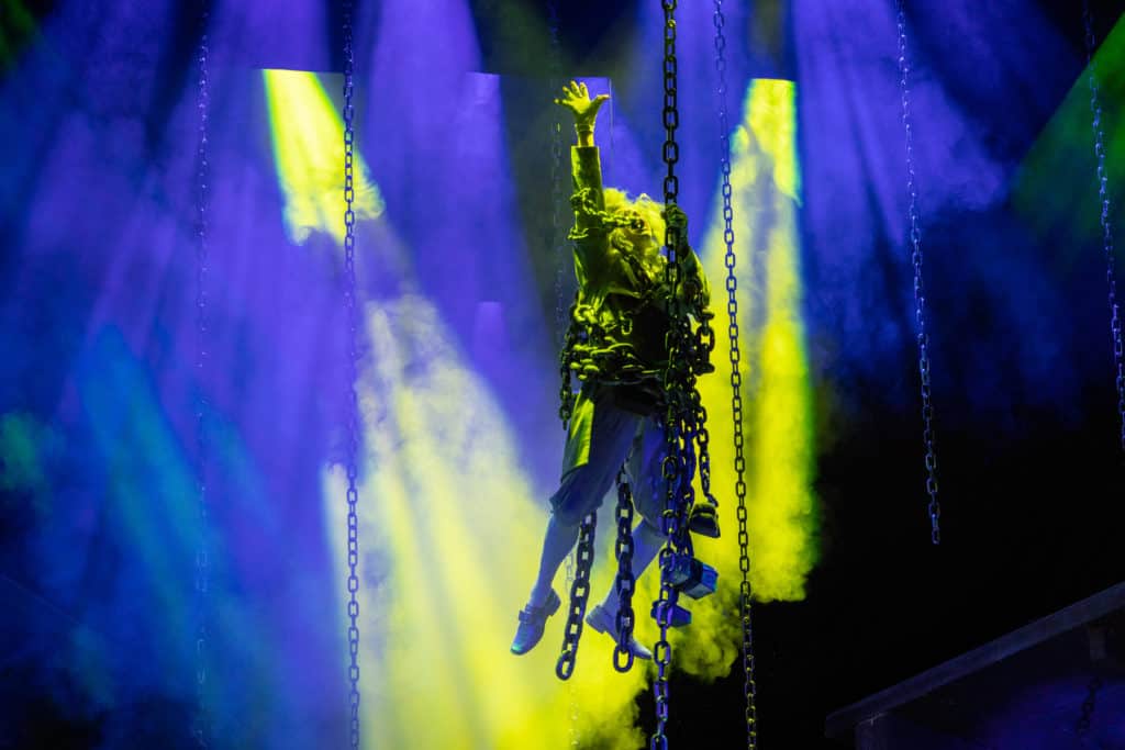 The Ghost of Jacob Marley descends in chains with smoke and blue and green colored lights behind him. He reaches one hand up in the air and the other hand holds the chain.