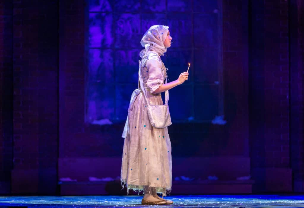 The Ghost of Christmas Past stands on stage holding a singular lit match. She is covered in white and stands alone on the stage in front of a blue-lit set.