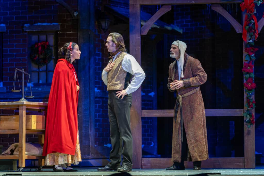 Belle and Young Ebenezer talk as present-day Ebenezer Scrooge looks on. Belle stands in a red cape and Young Ebenezer holds his hand on his chest.