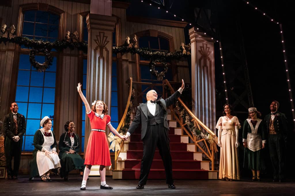 Annie and Daddy Warbucks performing on stage. Annie wears her classic red dress and her dad wears a tuxedo. They are standing in front of a grand staircase.