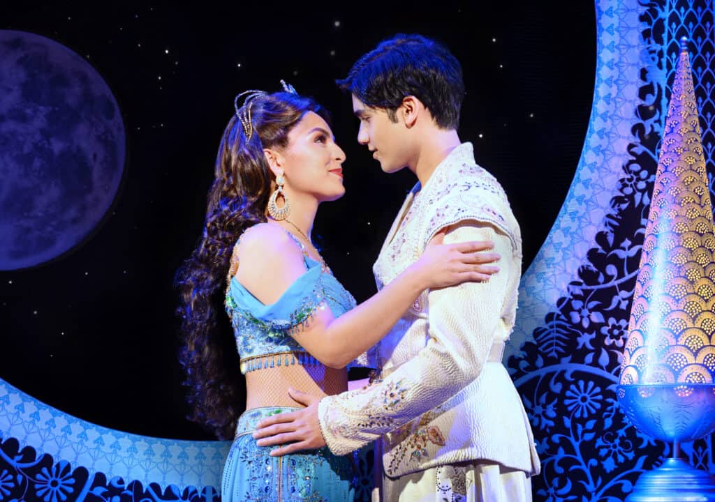 Aladdin and Jasmine stand in front of a blue backdrop and embrace.