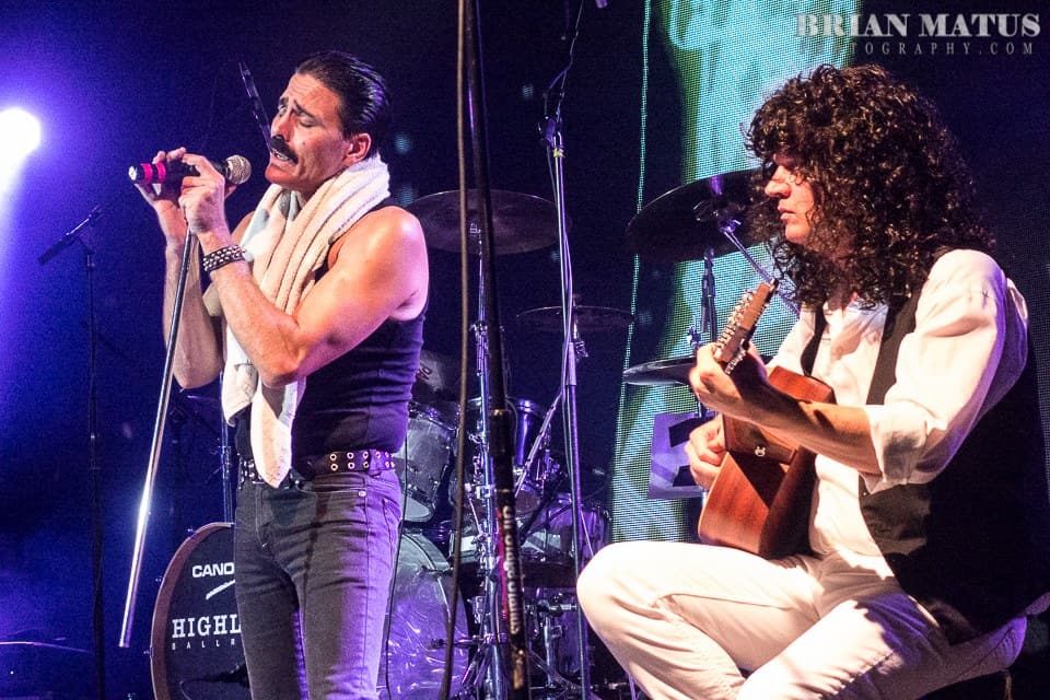 A Queen tribute band performs on stage. Joseph Russo as Freddie Mercury has a towel around his neck, and Steve Leonard as Brian May plays the guitar.