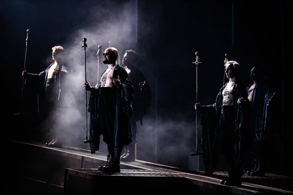 People with staphs stand on stage and are surrounded by smoke and dark lighting. 