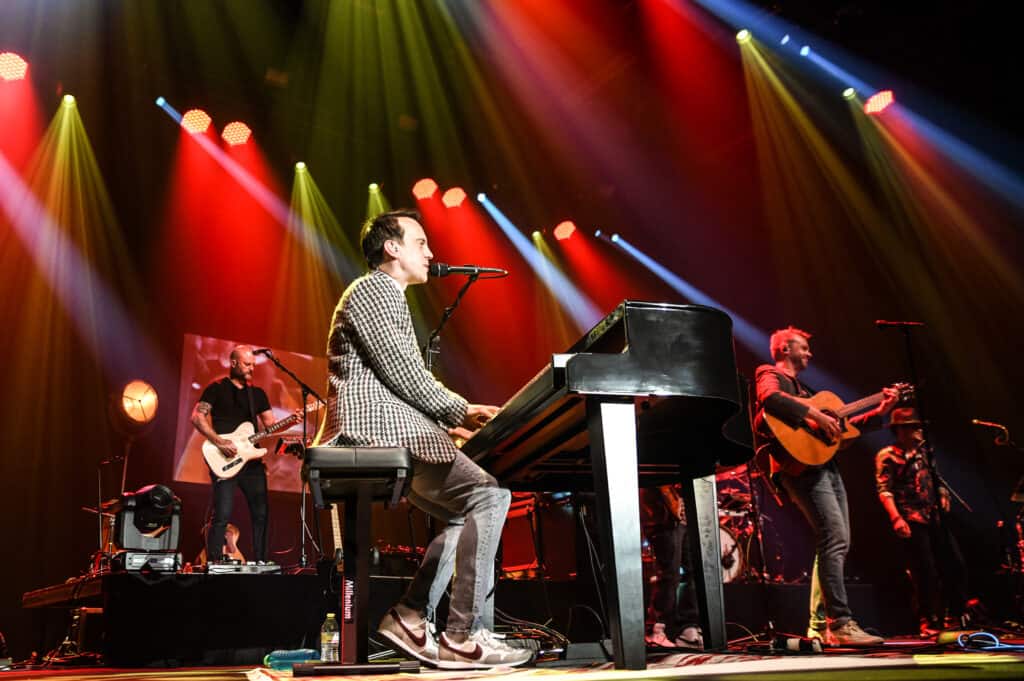 In front of red lights, Celebrating Billy Joel performs on stage. Rob Stringer plays the piano in a tweed suit.