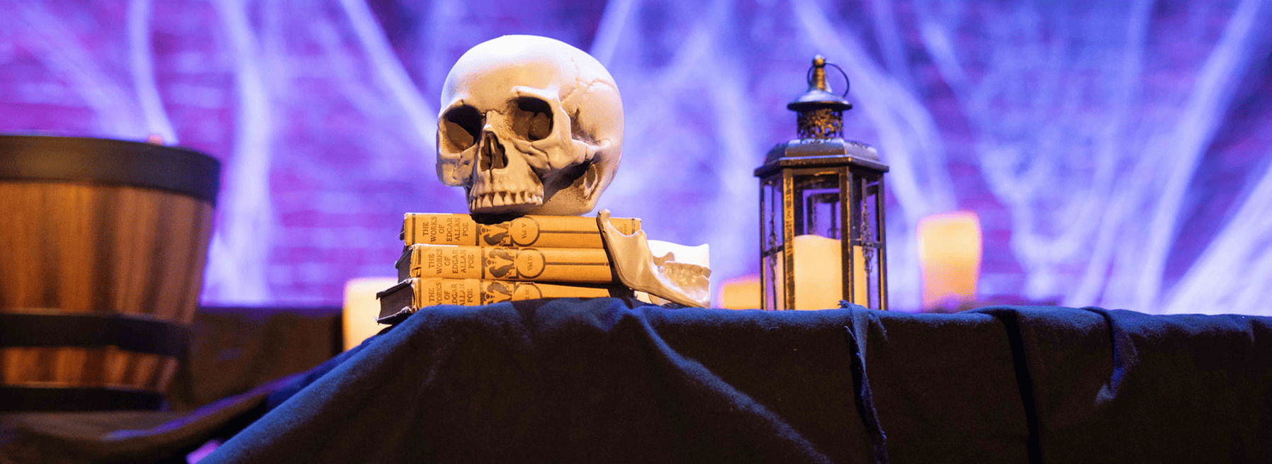 A skull prop sits atop three books next to a lantern. Spiderwebs adorn a brick wall in the background cast in purple-blue light