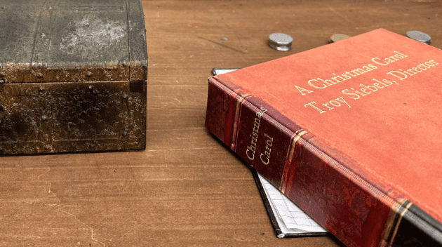 A leather bound binder reading "A Christmas Carol, Troy Siebels, Director" sits on a desk, closed.