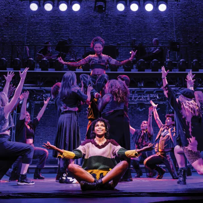 A number from Jagged Little Pill with one actor sitting crisscross and illuminated with a white spotlight front and center. Behind them in purple lighting are numerous dancers looking up at one being lifted above the rest.