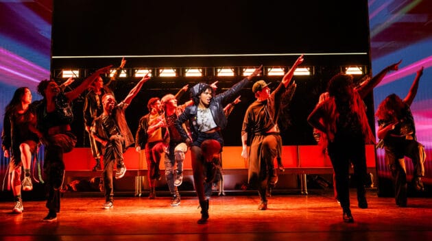 Teralin Jones and the Company of the North American Tour of JAGGED LITTLE PILL in a number where everyone on stage is dancing with one foot up and one hand pointing upwards. Front and center is the only actor in a white spotlight while everyone else is under red lights.