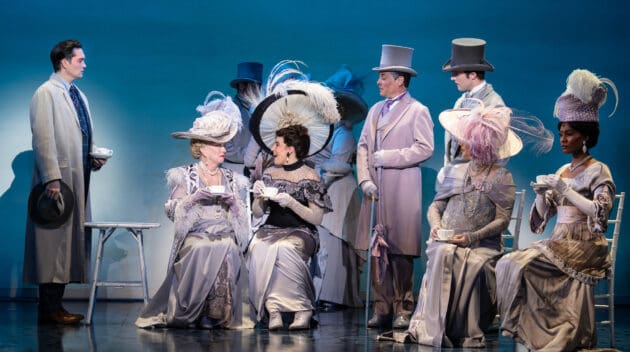 A tea party conversation in My Fair Lady. Four out of the six ladies are seated while the other two stand in the background and chat with a gentleman. Three other gentlemen stand around the seated ladies.