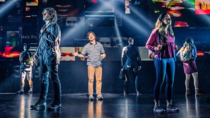 Evan from Dear Evan Hansen, a teen boy wearing a blue striped shirt, khakis and a cast on his forearm, is standing in the middle of a stage singing. Five people are standing around him with their backs turned to him as he sings.