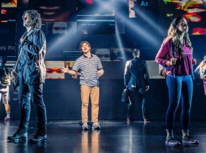 Evan from Dear Evan Hansen, a teen boy wearing a blue striped shirt, khakis and a cast on his forearm, is standing in the middle of a stage singing. Five people are standing around him with their backs turned to him as he sings.