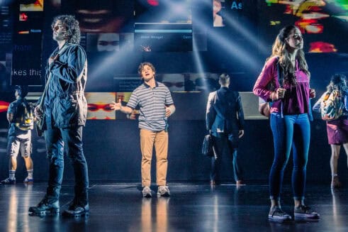 Evan from Dear Evan Hansen, a teen boy wearing a blue striped shirt, khakis and a cast on his forearm, is standing in the middle of a stage singing. Five people are standing around him with their backs turned to him as he sings.