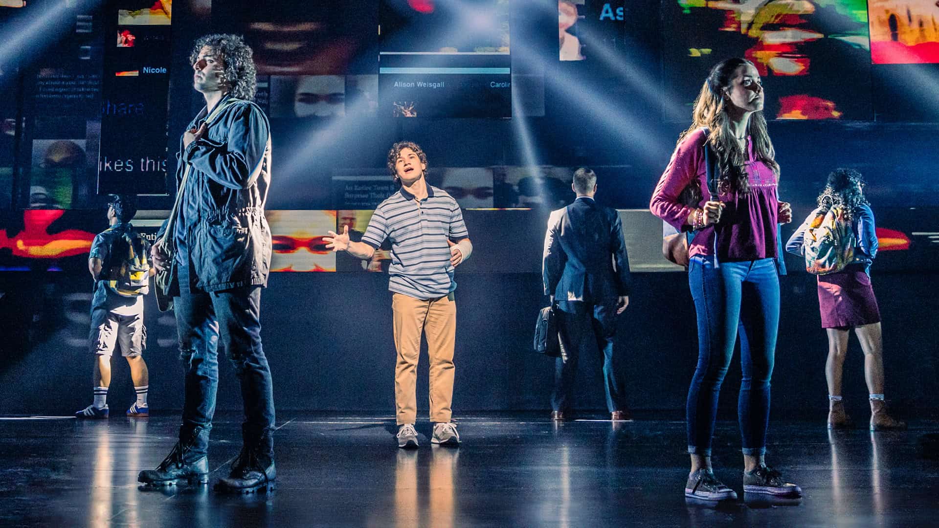 Evan from Dear Evan Hansen, a teen boy wearing a blue striped shirt, khakis and a cast on his forearm, is standing in the middle of a stage singing. Five people are standing around him with their backs turned to him as he sings.